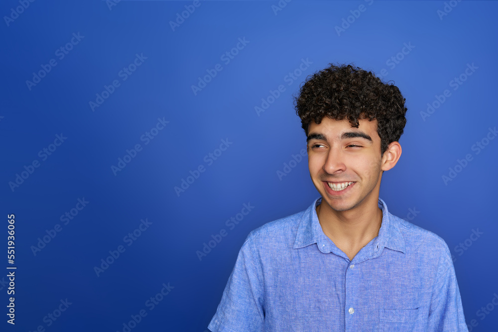 Funny young male with curly hair smiling while making face and rolling eyeballs against blue background Funny young male with curly hair smiling while making face and rolling eyeballs against blue background