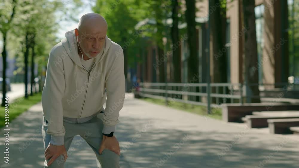 Mature male runner stopping to take deep breath during morning run on street wearing headphones. Sport, recreation and healthy lifestyle concept. Aging Youthfully Mature male runner stopping to take deep breath during morning run on street wearing headphones. Sport, recreation and healthy lifestyle concept. Aging Youthfully