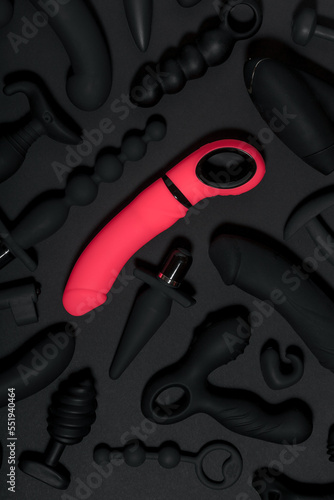 Erotic pleasure toys on a black background. Sex gadgets and masturbation devices. Pink dildo surrounded by black rubber and silicone sex toys: anal butt plugs, vibrators, vaginal balls. Top view.