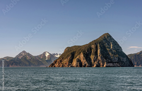 Resurrection Bay, Alaska, USA - July 22, 2011: Landscape with 1 isolated green forested triangular Hive Island in blue ocean water under light blue sky. Belt of snow patched mountains on horizon © Klodien