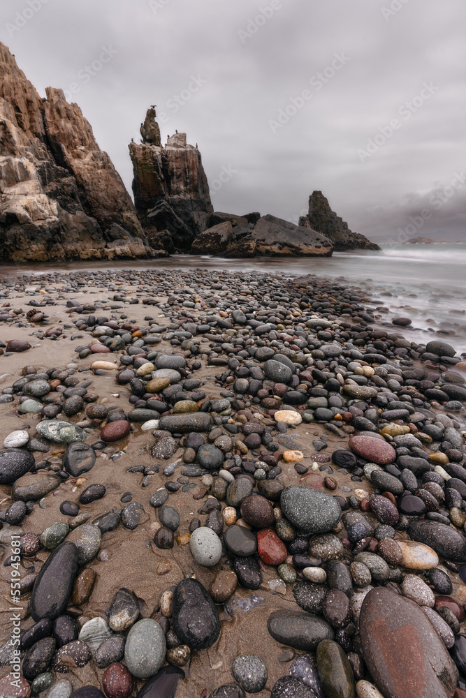 Vertical shot of sandy beach with stones, under a cliff, during a cloudy day.