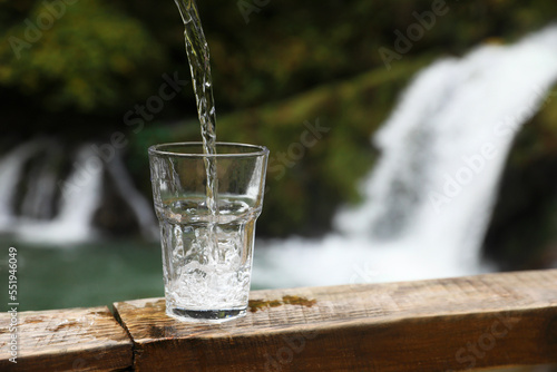 Fresh water pouring into glass on wooden surface near waterfall. Space for text