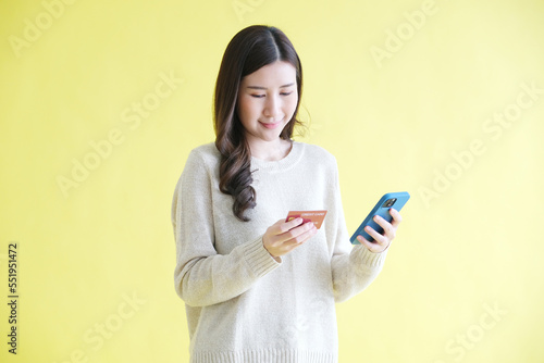 Young asian woman holding mobile phone and credit card for shopping online while standing over isolated yellow background