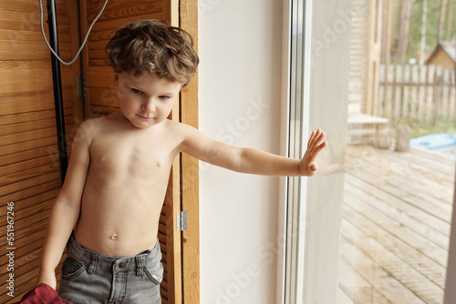 Little kid standing shirtless resting against window horizontal medium portrait