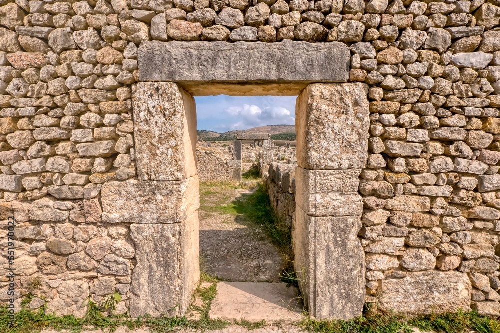 Looking through a doorway in a restored stone wall in the ancient former Roman city of Volubilis in northern Morocco, with a view of more ruins and the hilly countryside in the background.