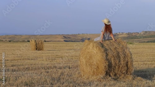 A beautiful young woman in a hat and a summer dress is sitting on a sheaf of hay in a field. Rural nature, wheat field, rest in the country, unity with nature