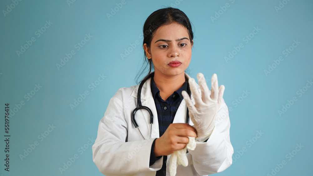 Portrait of a young female doctor wearing hand gloves, Asian Indian woman doctor in apron and stethoscope isolated over blue studio background
