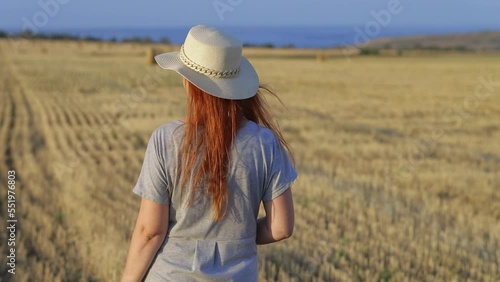 A beautiful young woman in a hat and a summer dress is sitting on a sheaf of hay in a field. Rural nature, wheat field, rest in the country, unity with nature