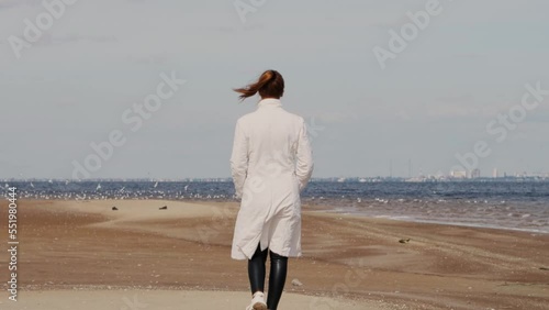 a beautiful woman with red hair in a white coat walks on a sandy beach on a cold sunny day