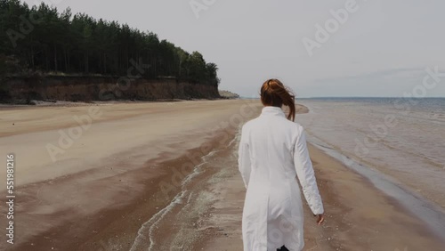 a beautiful woman with red hair in a white coat walks on a sandy beach on a cold sunny day