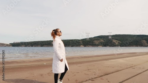 a beautiful woman with red hair in a white coat walks on a sandy beach on a cold sunny day