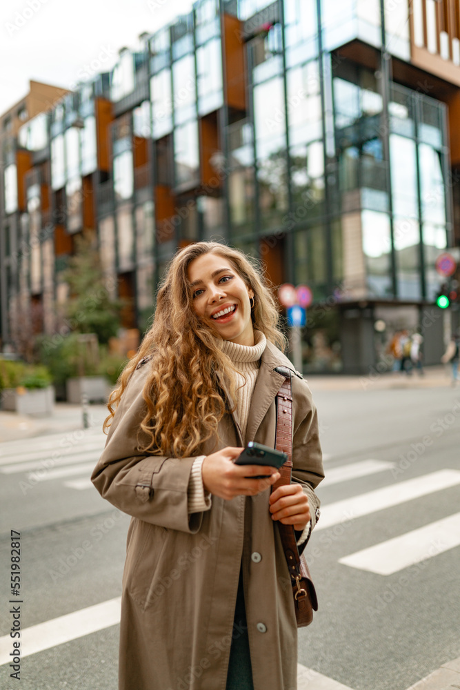 Fototapeta premium Smiling curly woman wearing warm coat walking down the street and using her phone
