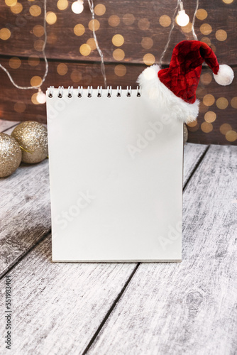 An empty notebook page with a Santa Claus hat, surrounded by Christmas decor and bokeh on a wooden background, copies the space. Christmas wish list, counting down the days
