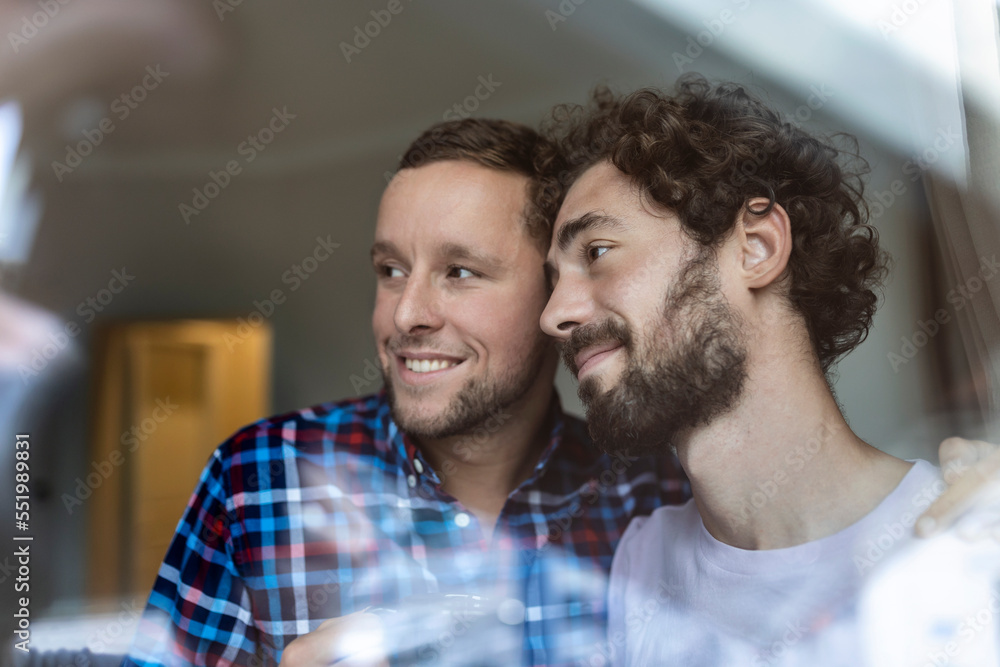 Fototapeta premium Young gay couple in love looking out the window. Two young androgynous men smiling together and having coffee.