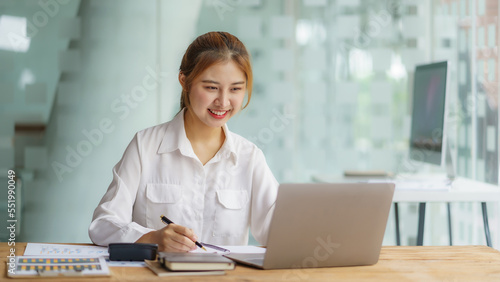 Young Asian businesswoman sitting happily working on her laptop and taking notes fluently and smiling happily at her assignment.