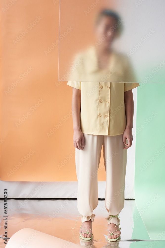 Full shot of young model hiding her upper body behind dim glass standing against orange, white and green background Full shot of young model hiding her upper body behind dim glass standing against orange, white and green background