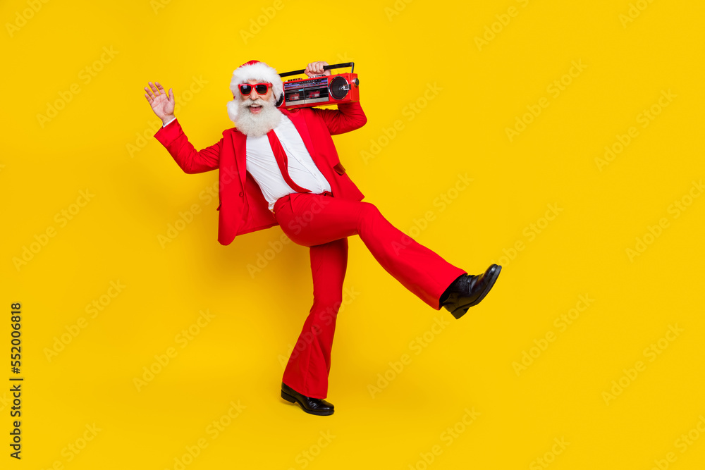 Full length photo of excited funky santa dressed red suit tie walking enjoying boom box music empty space isolated yellow color background Full length photo of excited funky santa dressed red suit tie walking enjoying boom box music empty space isolated yellow color background