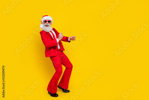 Full length photo of handsome pretty man pensioner dressed red suit santa hat having fun empty space isolated yellow color background