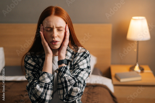 Portrait of sick redhead young woman with closed eyes touch head temples, suffering from headache, sitting on bed. Young pretty lady feeling unhealthy, strong sudden pain, morning hangover.