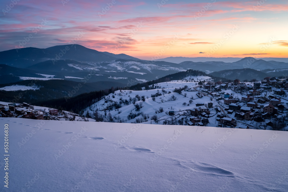 Beautiful sunrise view with snowy mountain slopes and small village among them in the frozen winter morning, the Rhodopi Mountains, Bulgaria