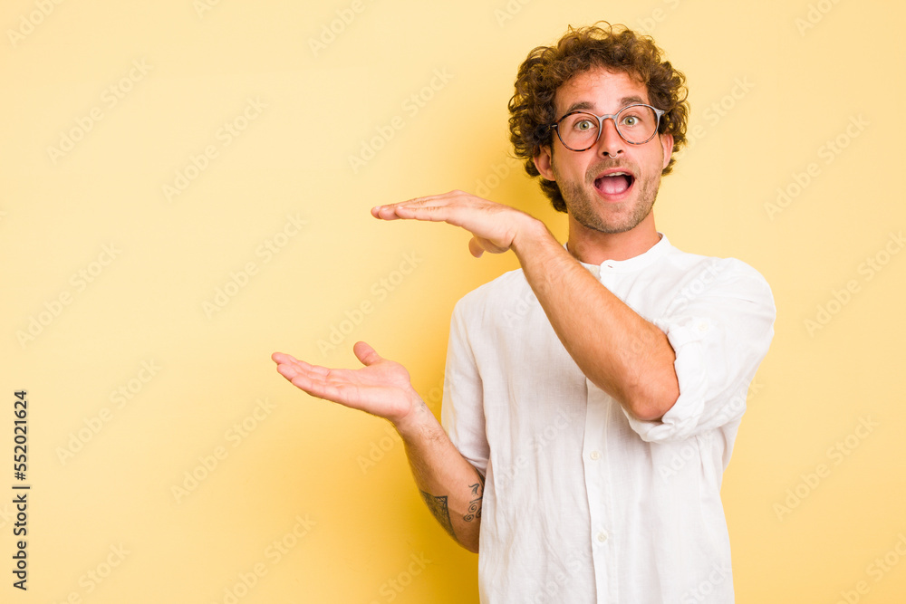 Young smart caucasian man on yellow background shocked and amazed holding a copy space between hands. Young smart caucasian man on yellow background shocked and amazed holding a copy space between hands.
