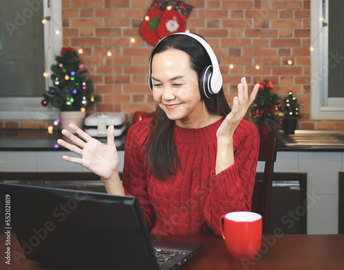 Asian woman wearing red knitted sweater and headphones sitting  with red cup  in the kitchen with Christmas decoration, using computer notebook video call talking with friends or family. photo