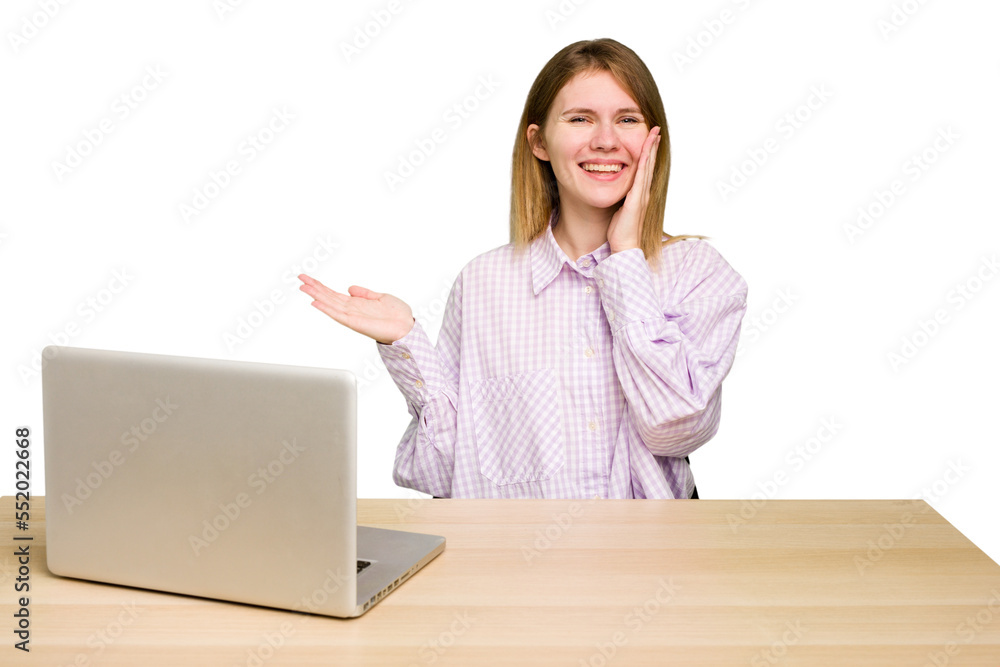 Young caucasian woman in a workplace working with a laptop isolated holds copy space on a palm, keep hand over cheek. Amazed and delighted. Young caucasian woman in a workplace working with a laptop isolated holds copy space on a palm, keep hand over cheek. Amazed and delighted.