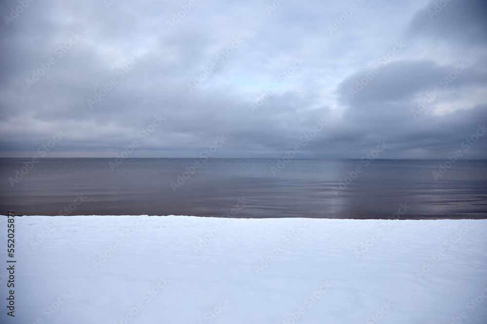 Baltic sea coast covered with white snow, calm sea water with grey clouds above, selective focus