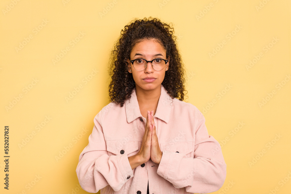 Young Brazilian curly hair cute woman isolated on yellow background praying, showing devotion, religious person looking for divine inspiration. Young Brazilian curly hair cute woman isolated on yellow background praying, showing devotion, religious person looking for divine inspiration.