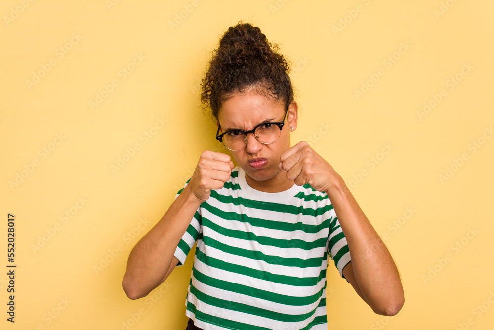Young Brazilian curly hair cute woman isolated on yellow background throwing a punch, anger, fighting due to an argument, boxing. Young Brazilian curly hair cute woman isolated on yellow background throwing a punch, anger, fighting due to an argument, boxing.