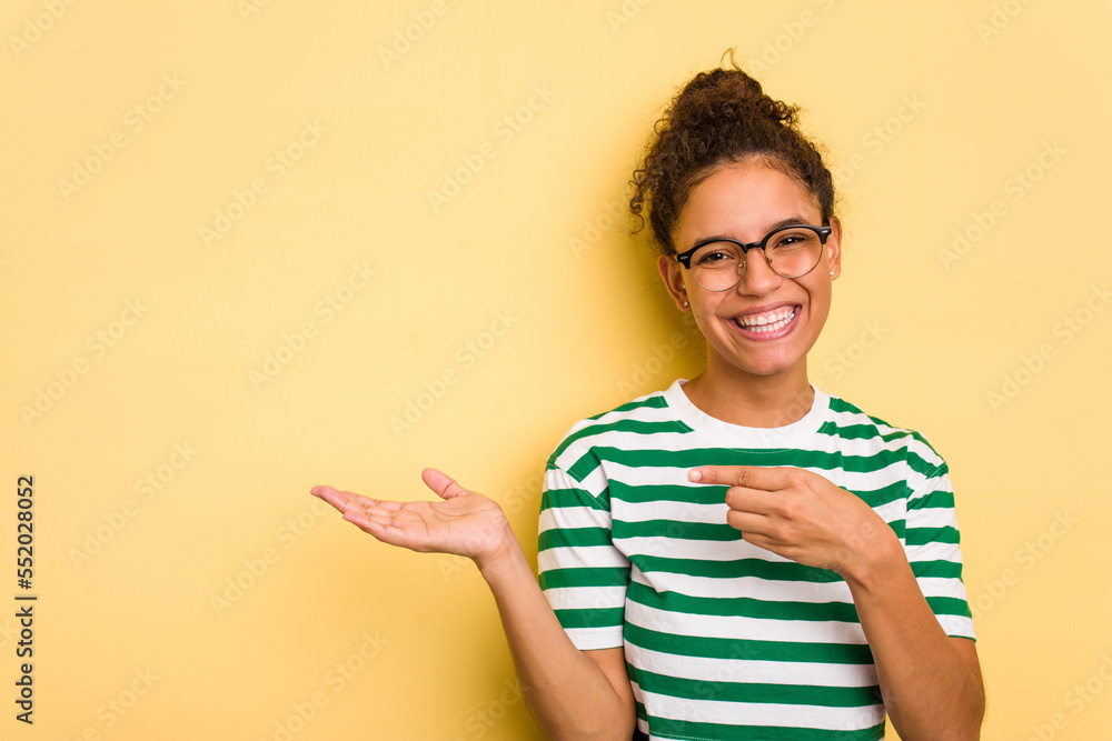 Young Brazilian curly hair cute woman isolated on yellow background excited holding a copy space on palm. Young Brazilian curly hair cute woman isolated on yellow background excited holding a copy space on palm.