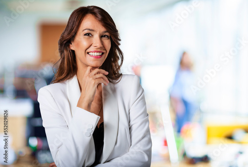 Portrait of happy brunette female looking at camera and laughing. Close-up of an attractive middle aged woman with toothy smile wearing blazer while standing inside at office. Copy space. 