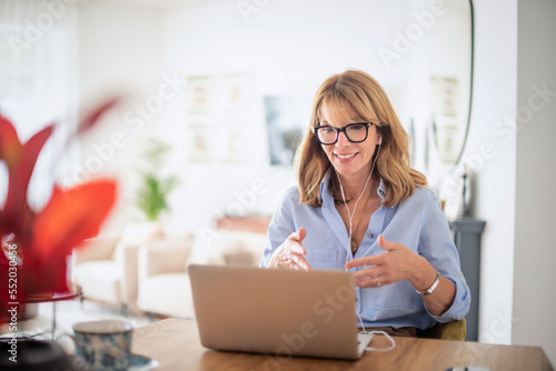 Middle aged woman sitting at her dining room table and using earphone and laptop while having video call. Home office.  photo