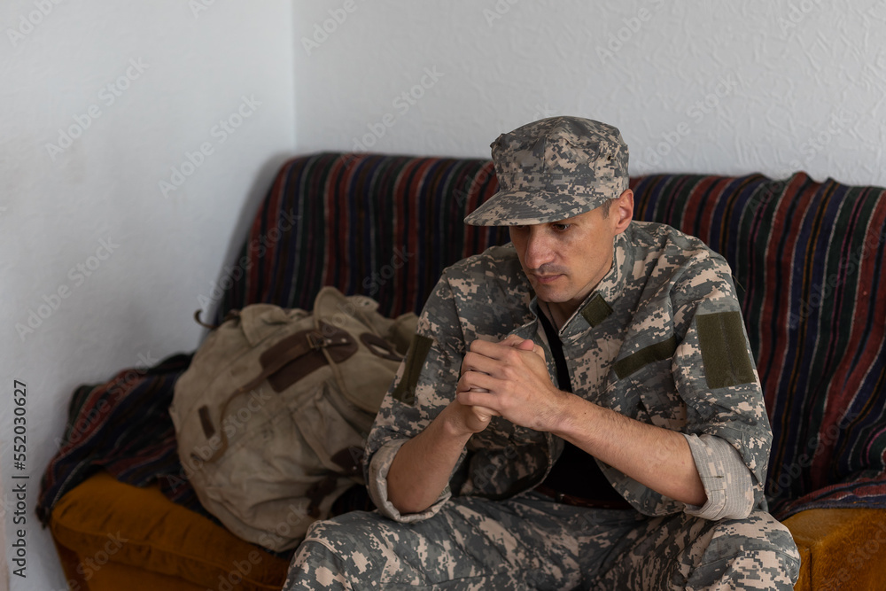 Unhappy man in camouflage uniform sitting at arm chair over white studio background, touching his head, soldier suffering from posttraumatic stress disorder, returning back home after military service