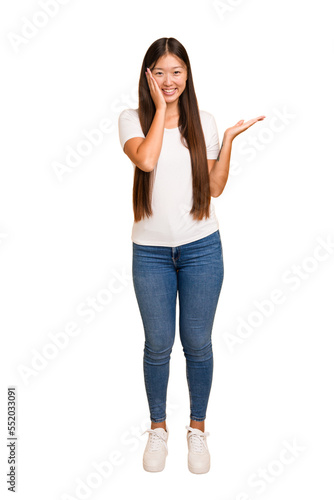 Young asian woman standing, full body cutout isolated holds copy space on a palm, keep hand over cheek. Amazed and delighted.