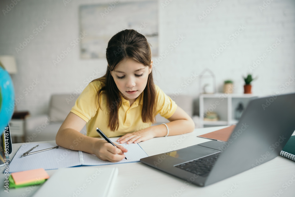 preteen girl writing in notebook while learning near laptop in living room preteen girl writing in notebook while learning near laptop in living room