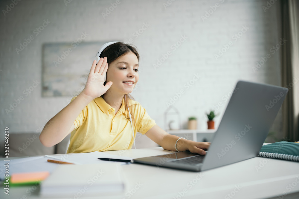 smiling girl in headphones waving hand near laptop during online lesson at home smiling girl in headphones waving hand near laptop during online lesson at home