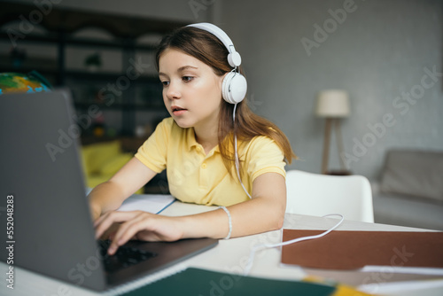 brunette schoolgirl in wired earphones learning at home and typing on laptop