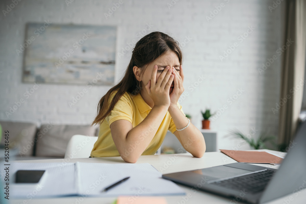 exhausted kid with closed eyes covering face with hands while sitting near blurred laptop at home exhausted kid with closed eyes covering face with hands while sitting near blurred laptop at home