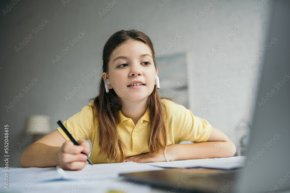smiling girl in wireless earphones holding pen and looking at laptop on blurred foreground smiling girl in wireless earphones holding pen and looking at laptop on blurred foreground