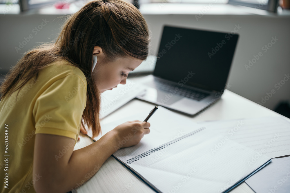 brunette girl in wireless earphone writing in notebook near blurred laptop at home brunette girl in wireless earphone writing in notebook near blurred laptop at home