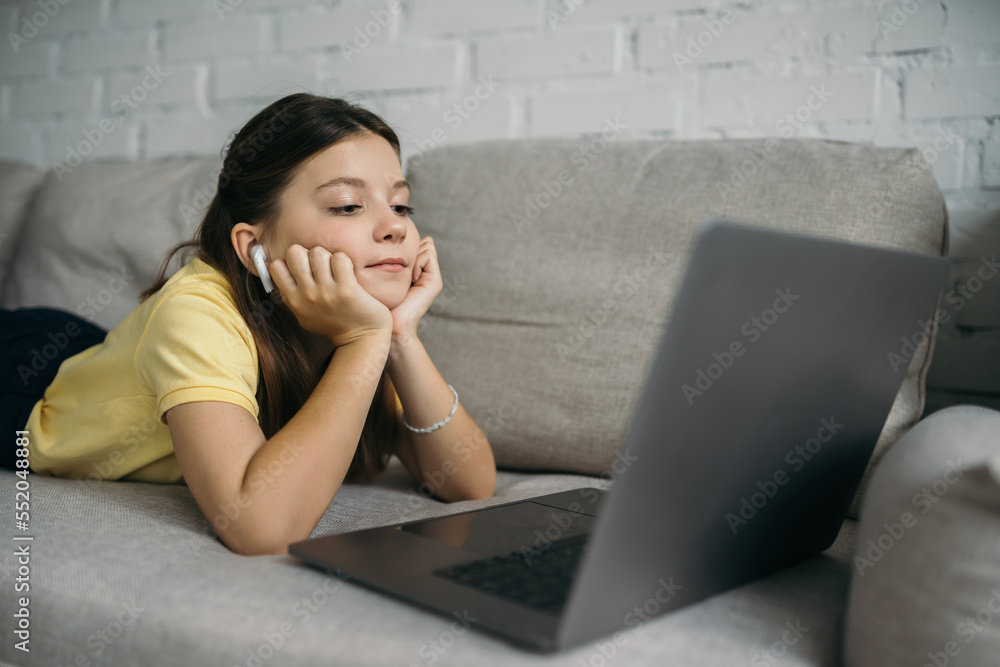 brunette girl in wireless earphone looking at blurred laptop while lying on cozy couch at home brunette girl in wireless earphone looking at blurred laptop while lying on cozy couch at home