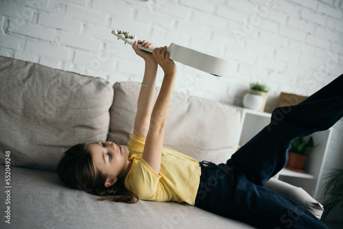preteen girl lying on couch in living room and holding ukulele in raised hands