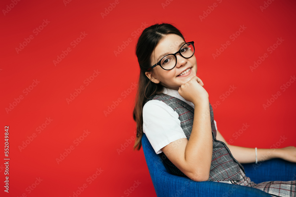pleased girl in trendy eyeglasses sitting in armchair with hand near chin isolated on red pleased girl in trendy eyeglasses sitting in armchair with hand near chin isolated on red