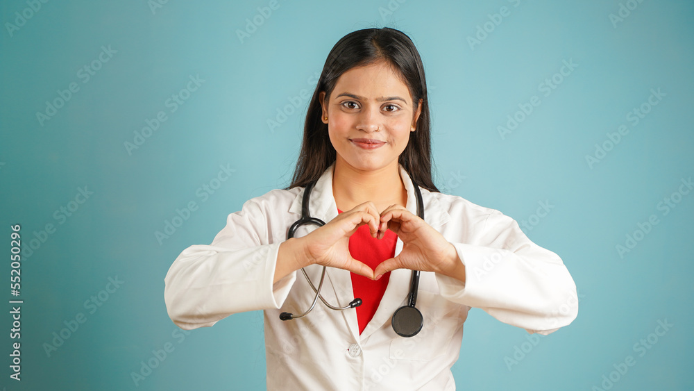 Portrait of a young female doctor cardiologist showing heart symbol with fingers, Cheerful Asian Indian woman doctor in apron and stethoscope isolated blue studio background, expressing love and care Portrait of a young female doctor cardiologist showing heart symbol with fingers, Cheerful Asian Indian woman doctor in apron and stethoscope isolated blue studio background, expressing love and care