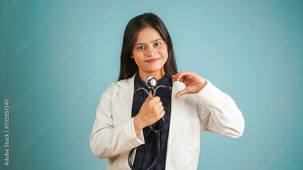 Portrait of a young female doctor cardiologist showing heart symbol with fingers, Cheerful Asian Indian woman doctor in apron and stethoscope isolated blue studio background, expressing love and care Portrait of a young female doctor cardiologist showing heart symbol with fingers, Cheerful Asian Indian woman doctor in apron and stethoscope isolated blue studio background, expressing love and care