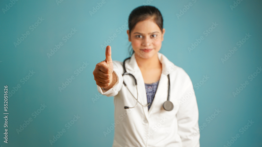 Portrait of a young female doctor gesturing thumbs up, Cheerful Asian Indian woman doctor in apron and stethoscope isolated over blue studio background Portrait of a young female doctor gesturing thumbs up, Cheerful Asian Indian woman doctor in apron and stethoscope isolated over blue studio background