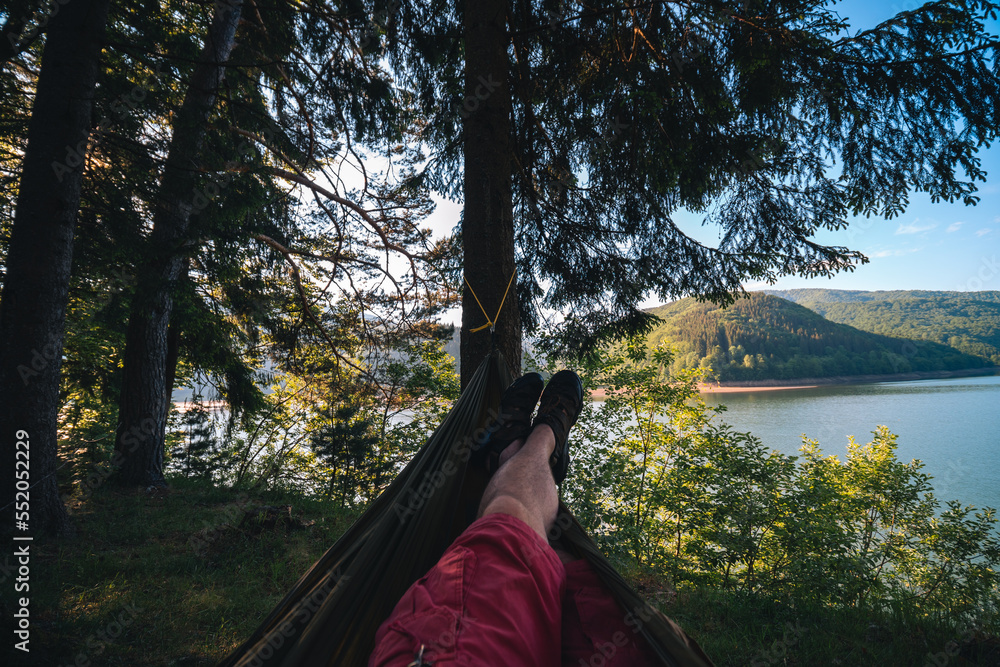 Man in hammock, first person look view, warm summer day, pine tree. river and mountains background. Travel and vacation, tourism equipment concept. Copy space