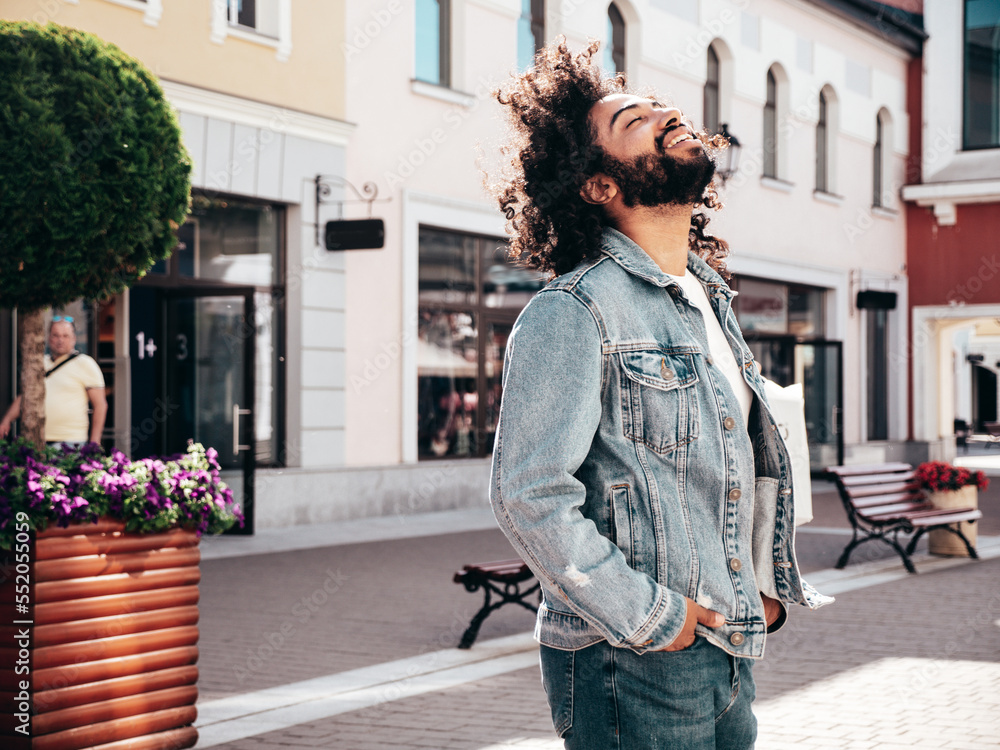 Handsome smiling hipster  model. Sexy unshaven Arabian man dressed in summer jeans jacket clothes. Fashion male with long curly hairstyle posing in street at sunset. Cheerful and happy