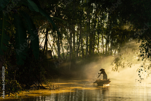 Photo of a local asian old man male boatman wearing conical hat rowing a small wooden boat across a small river during sunset time in a bamboo forest to deliver some dry grasses as animal feeds. 